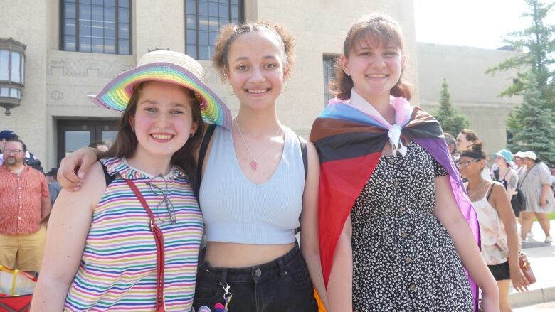 Three teenage girls are standing and smiling at the camera. One is wearing a rainbow-coloured wide-brim hat, another has a Pride flag draped over her.