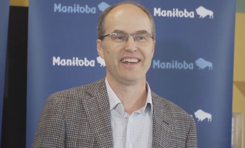 A bald man with glasses and a checkered suit speaks in front of a backdrop that has the word Manitoba on it.