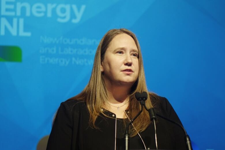 A woman with long brown hair in a black suit delivers a speech.