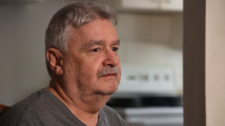 A man with a moustache and wearing a grey t-shirt sits in an apartment.