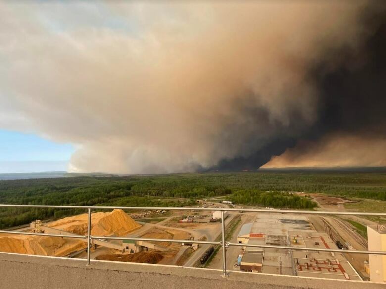 A photo from a high industrial building overlooks a pulp mill and a wildfire generating a huge plume of smoke above the forest in the distance.