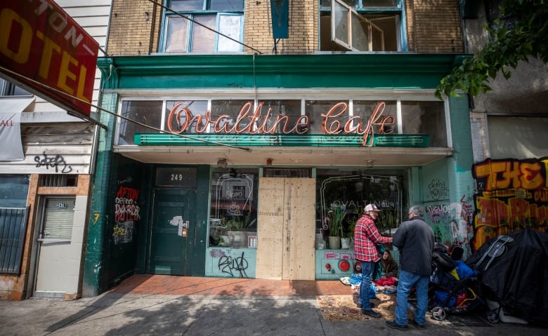 The Ovaltine Cafe signage is centred above a closed storefront plastered with graffiti. Two men stand, and a woman sits on a mat outside next to a tent and a pile of objects. 