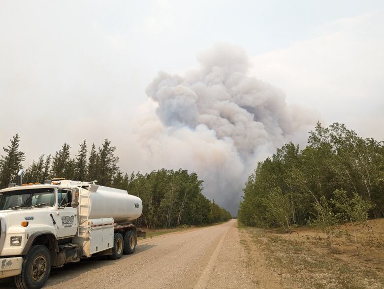 A tanker truck is parked on a dirt road, with wildfire smoke visible in the background.