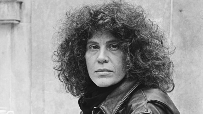 A black and white portrait of a woman with curly black hair and a black leather jacket looking over her shoulder into the camera.