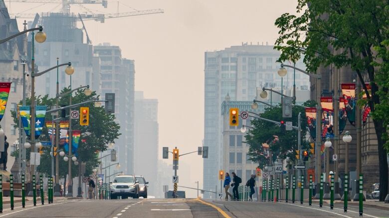 People walk across an intersection with buildings behind them obscured by smoky sky due to wildfires