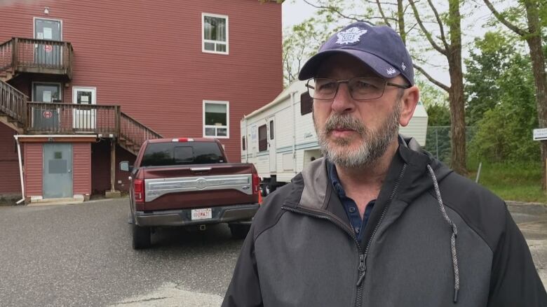 A man with facial hair wears a grey jacket and a Toronto Maple Leads hat. He stands in front of a red house.