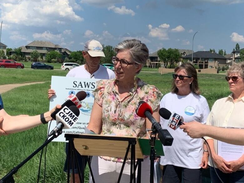 A woman in a floral shirt speaks into microphones surrounded my people, some with signs. 