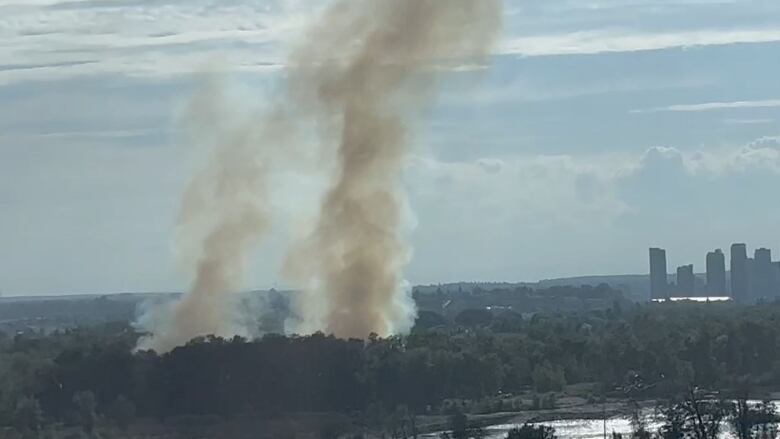 Smoke rises from a park area near a river with the city skyline on the right.