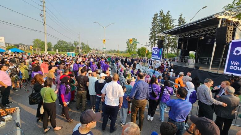 A crowd gathers in front of a stage set up on a roadway. The stage is flanked by signs reading 'We remember #OurLondonFamily.'