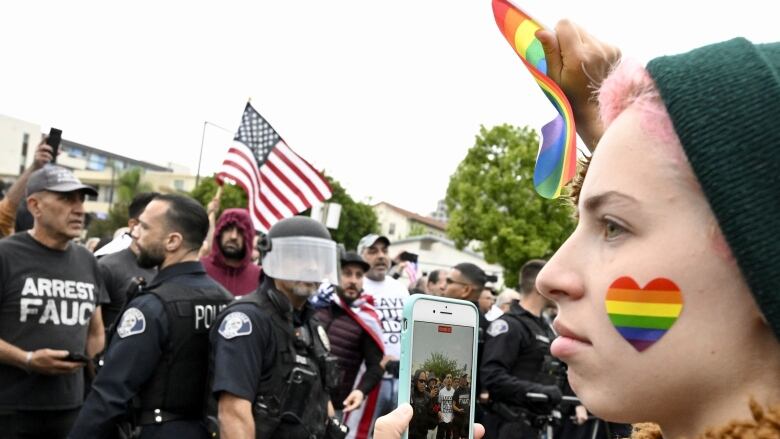 A person on the right side of the frame has a rainbow-striped heart on their face and holds a small rainbow flag in the air in front of police blocking a row of protesters on the left side of the image. 