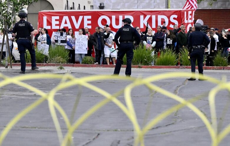 Three police officers stand in a parking lot facing a line of protesters holding placards in front of a large, red banner with the words 