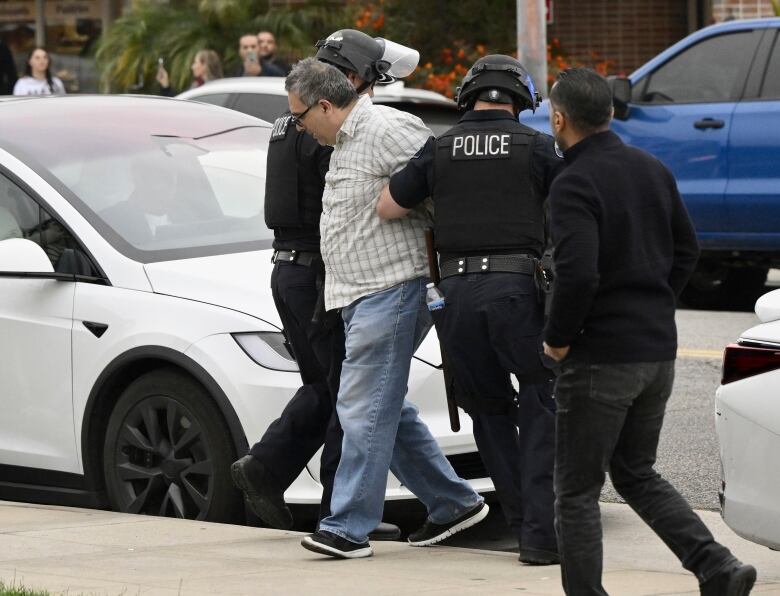 Two uniformed police officers walk a man past a white car. 