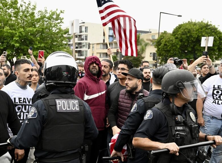 Two police officers, wearing helmets and holding batons,  stand in front of a crowd of men.