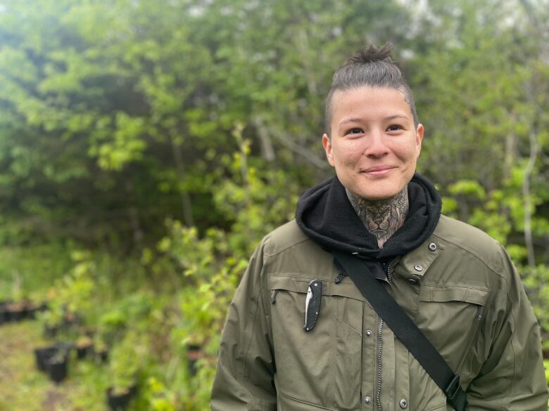 Em McFadyen stands against a forested backdrop. They have a tattoo of a lion along their throat with hair in a top knot. 