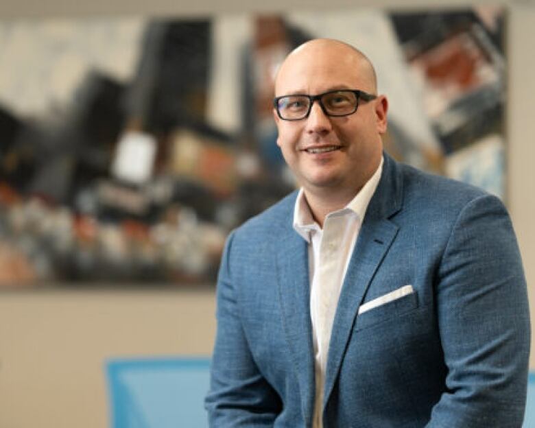 A bald man in glasses and a suite smiles and sits on a desk in an office.