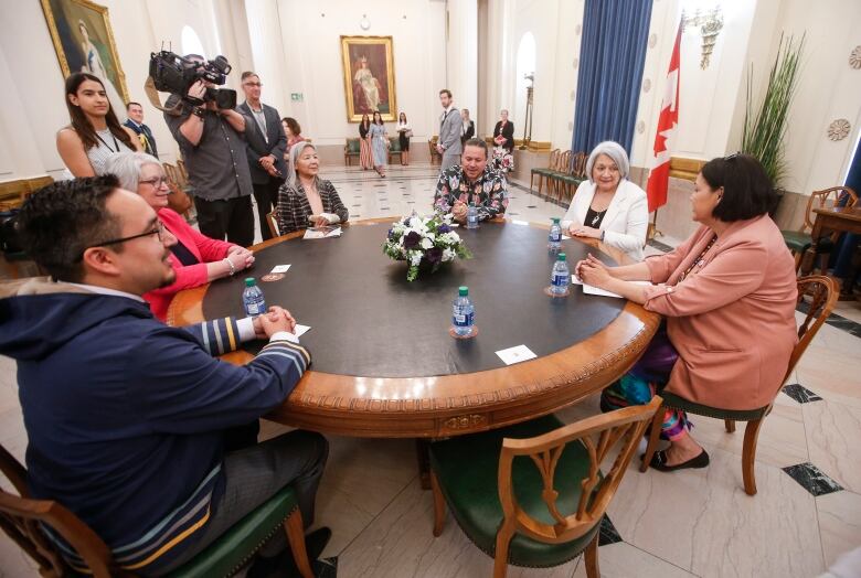 Several people, including a white-haired woman in a white suit jacket, sit at a round table, with members of the news media standing in the background.