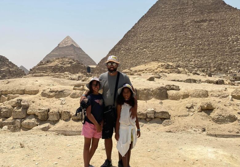 A man and two two dressed in shorts and T-shirts pose in front of pyramids amid a sandy and rocky background. 