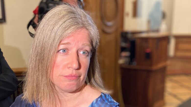 A woman with blond hair, wearing a blue top glances over her left shoulder as a man in a suit on her right holds a tape recorder near her. 