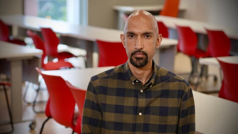 A bald man with a goatee sits in a classroom with red chairs.