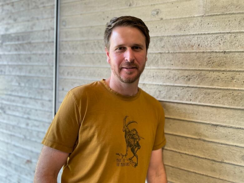 A man with beard and in yellow T-shirt stands in front of a wall.