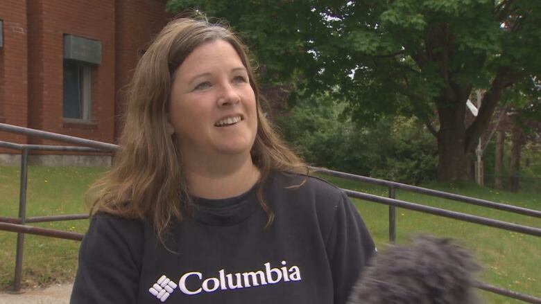 A woman with shoulder length hair in a black sweatshirt speaks to the camera.