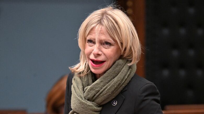 A blond woman wearing a green scarf and black blazer speaks at the National Assembly.