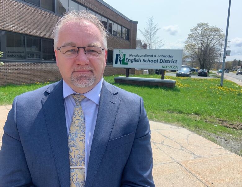 A person wearing a suit and tie stands in front of a sign with the Newfoundland and Labrador English School District logo.