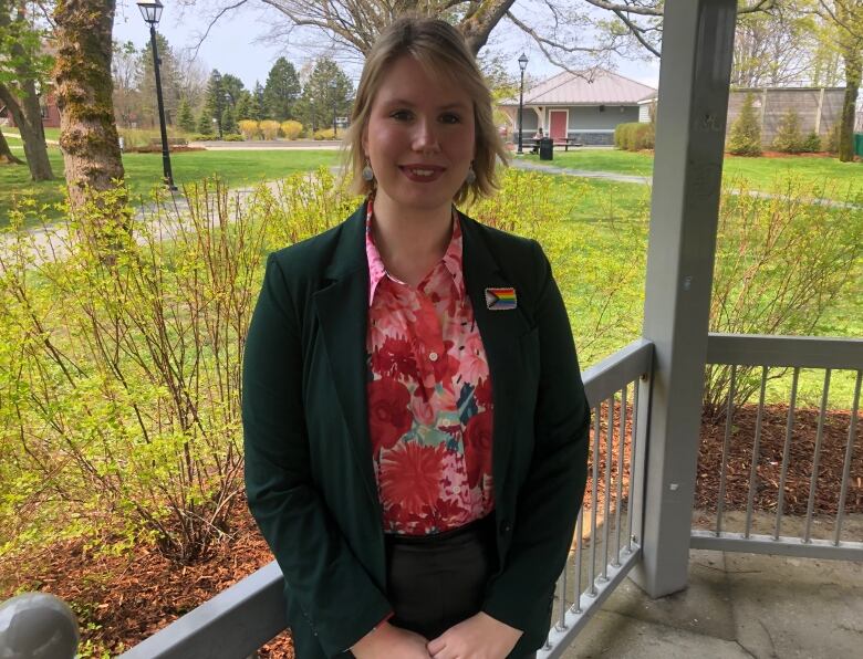 A person wearing a blazer with a Pride flag pinned to it stands under a gazebo.