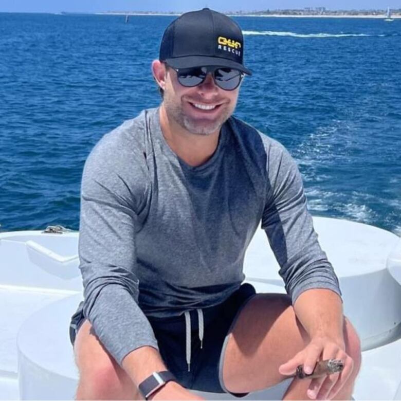 A man wearing sunglasses and a ball cap in swim trunks and a sweater sits in a boat on a white bench, smoking a cigar and smiling broadly at the camera while the vast expanse of a deep blue sea is laid out behind him.