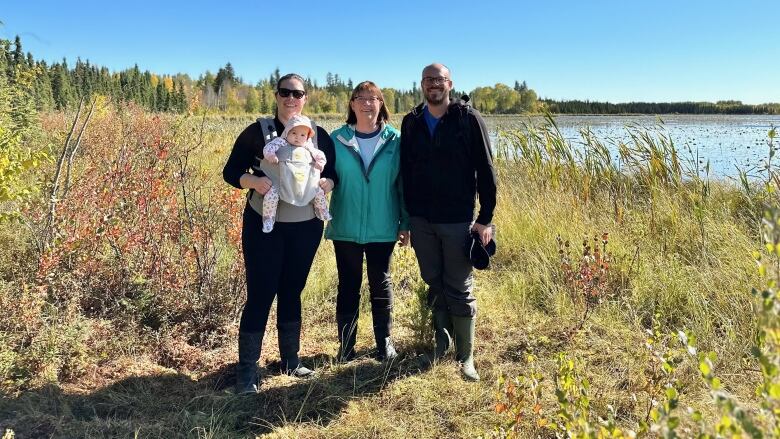 A woman, holding a baby, a mother, and a man stand in rubber boots at the edge of a lake at JBJ McDonald Conservation Land, west of Edmonton. 