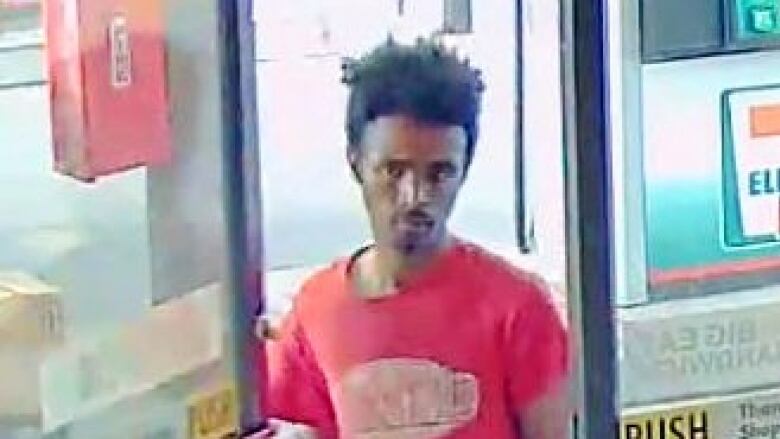 A man opens the door of a convenience store. He is wearing a red shirt. 