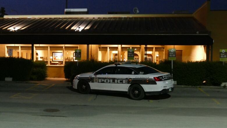 A black and white police cruiser sits parked in front of the windows of a restaurant at night.