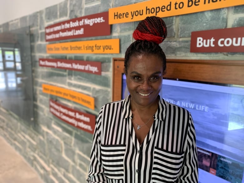 A woman wearing a black-and-white striped shirt smiles as she stands in front of a stone wall adorned with signs and a television.