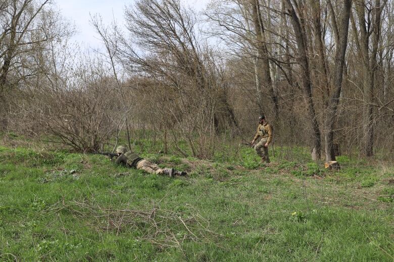 A person standing at a distance watches over another person lying on the ground and pointing a military weapon.