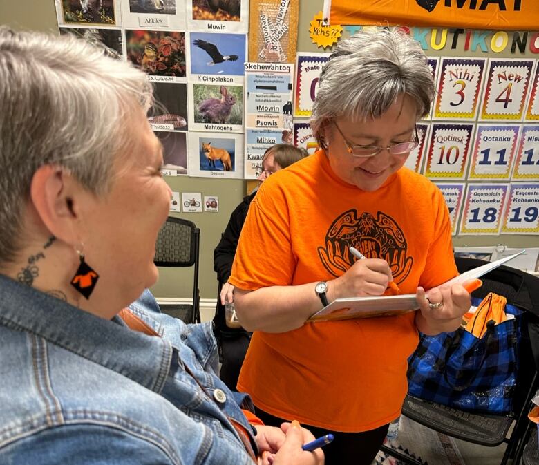 A woman wearing an orange shirt signs a book for a woman wearing a denim jacket.