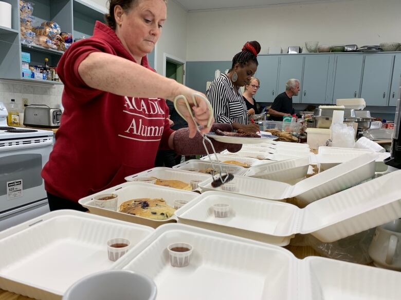 A woman uses metal tongs to put pancakes and sausages into cardboard takeaway containers. More volunteers are prepping other meals in the background.