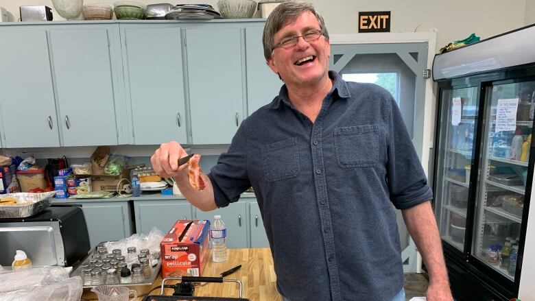 A man stands next to a griddle full of bacon. He's holding up a piece of bacon with tongs and smiling.