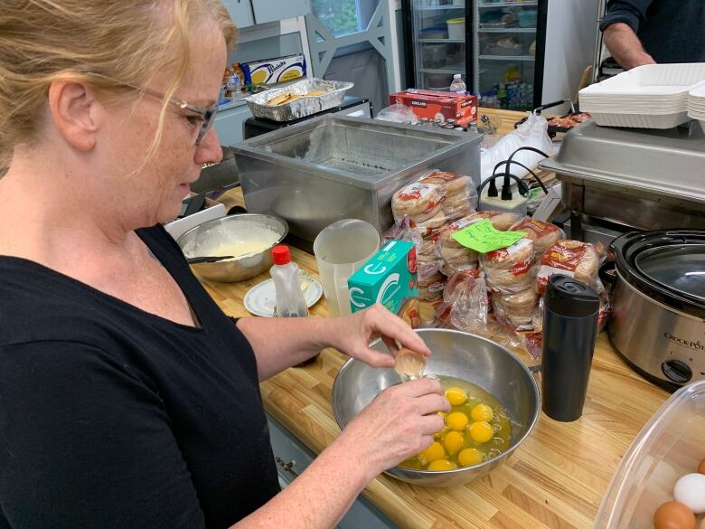 A woman in a black t-shirt is cracking eggs into a metal bowl. There are stacks of other breakfast ingredients on the counter around her.