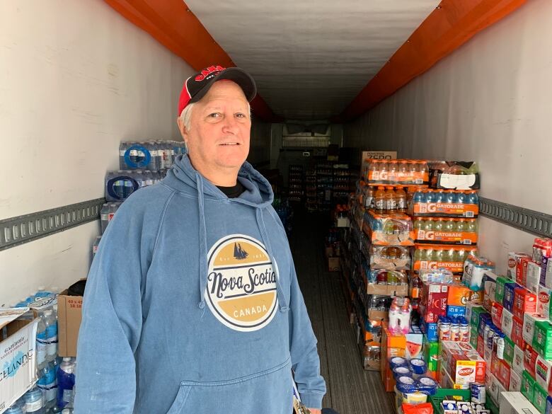 A man in a blue Nova Scotia hoodie stands in the back of a reefer truck. There are flats of soda, water bottles, and other beverages stacked behind him.