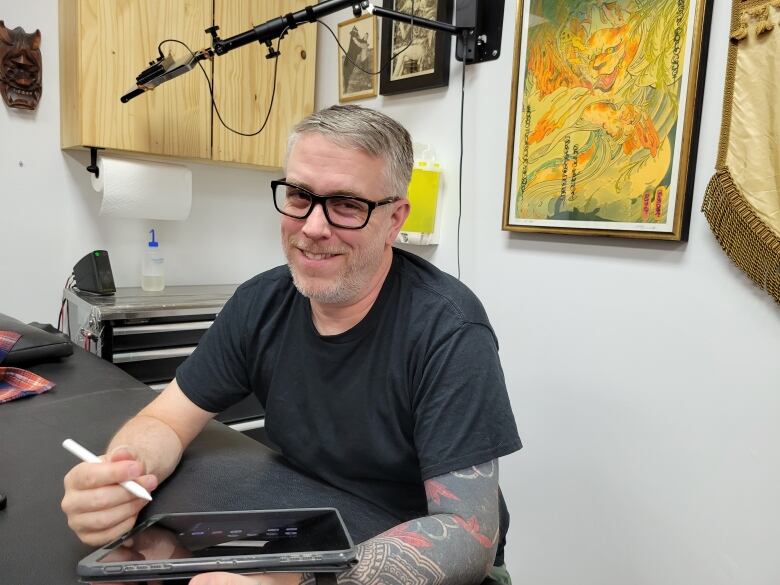  A tattooed man sits with a smile in an office holding a tablet.