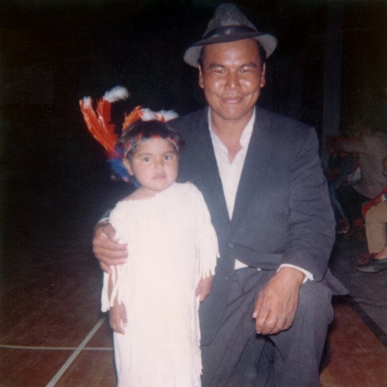 A little girl in a light coloured dress stands beside her kneeling father in a suit smiling at the camera. 