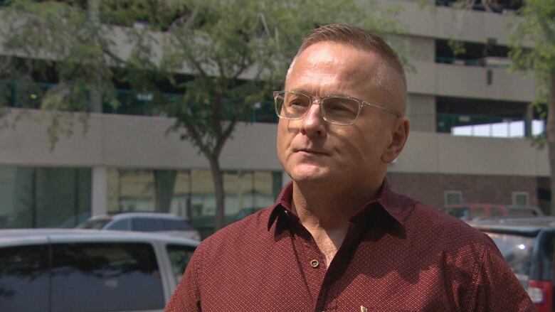 man in red button-up shirt stands outside with a parkade behind him