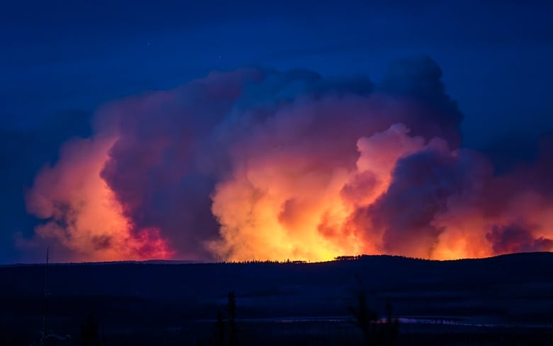 A wildfire is shown burning on a hillside at night, with smoke billowing into the air.