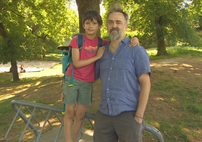 A young boy with a red shirt and blue backpack stands on a bike rack. A man in a blue button up shirt holds him with one arm and his other hand in his pant pocket. 