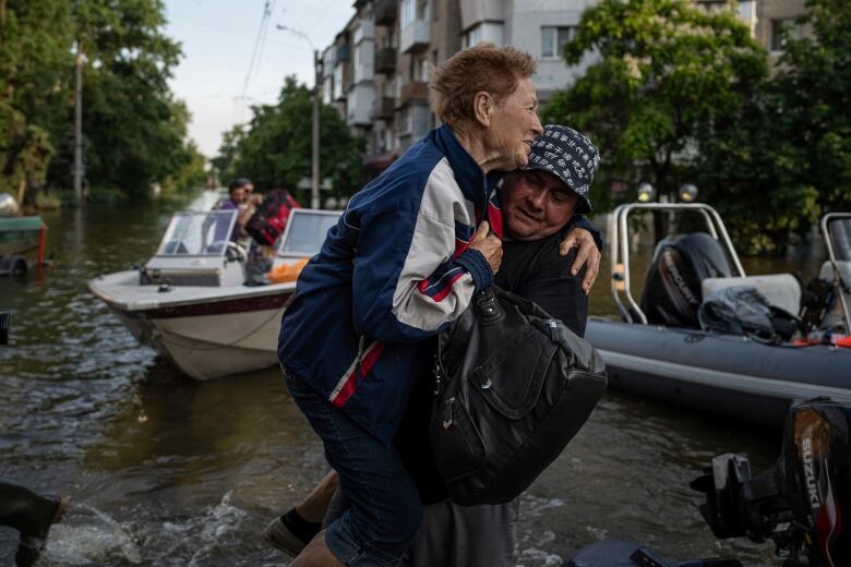 A man hoists a woman as he walks in water. Two small boats and a building can be seen in the background.