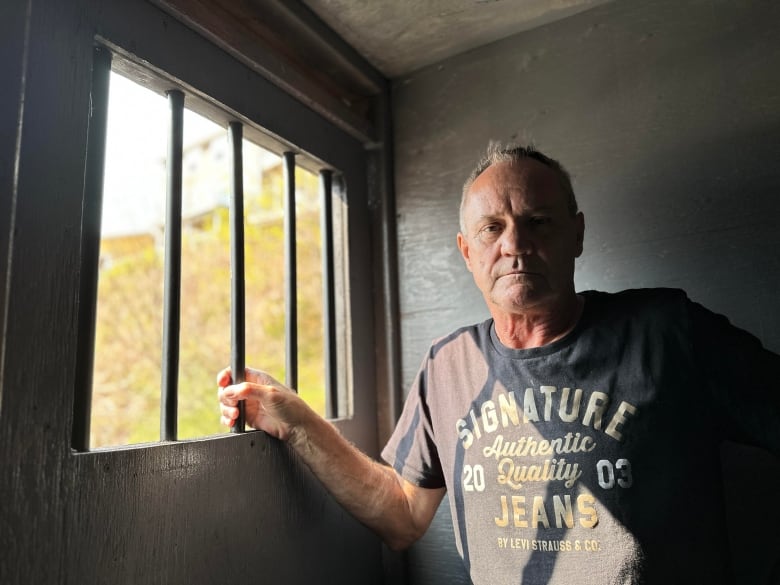 A man stands inside a makeshift jail cell, with one hand wrapped around the bars on the window.