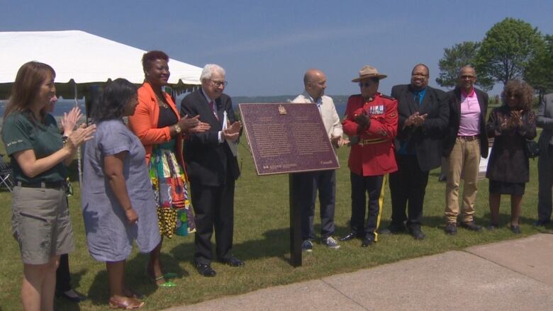 A group of nine people stand around a dark red plaque. They are applauding, as it was just unveiled. 