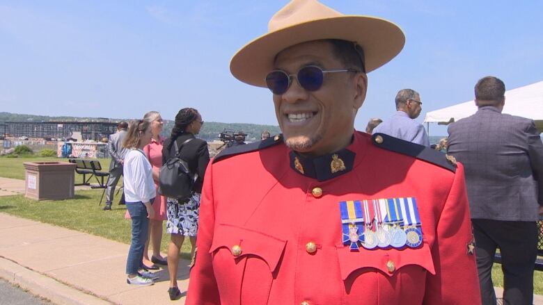 A main is wearing his RCMP uniform. He wears a red coat with medals on his chest. He's smiling, wearing sunglasses and a tan hat. 