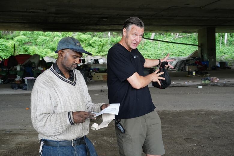 Jacco Stuben, left, looks at his tax return, one of the documents he needs to get a subsidized apartment. David Chapman from Resilience Montreal stands to his right motioning toward the tent community behind them.  