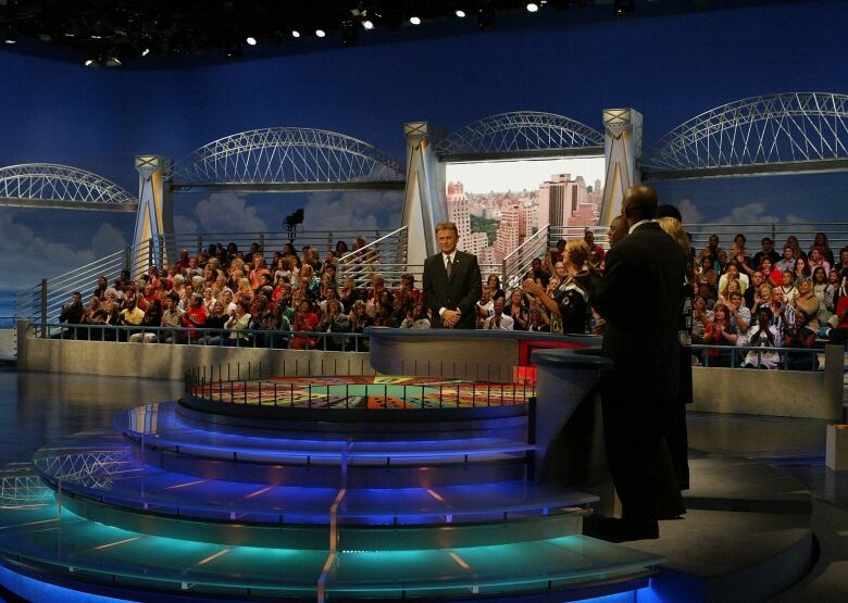 A host and contestants are seen onstage of a game show as studio audience members sit and watch in the background.